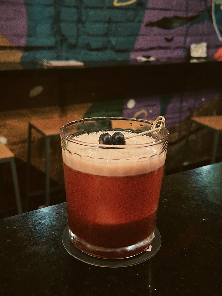 Uma mão segura um copo de chope claro na frente da parede de tijolos pintada com o mural roxo, verde e vermelho do bar.