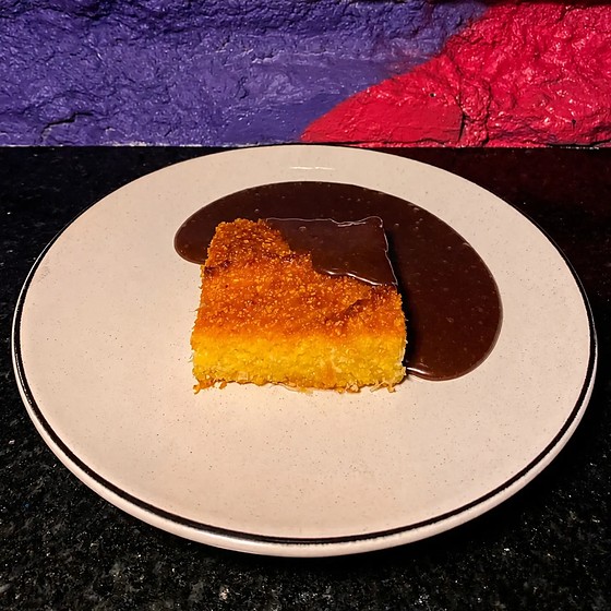 Fatia de bolo de milho com coco num prato claro, ao lado de uma poça de ganache de chocolate.