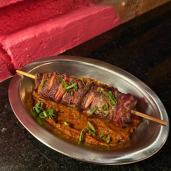 Espetinho de gado e bacon assado na brasa, numa travessa de metal com molho vermelho por baixo.
