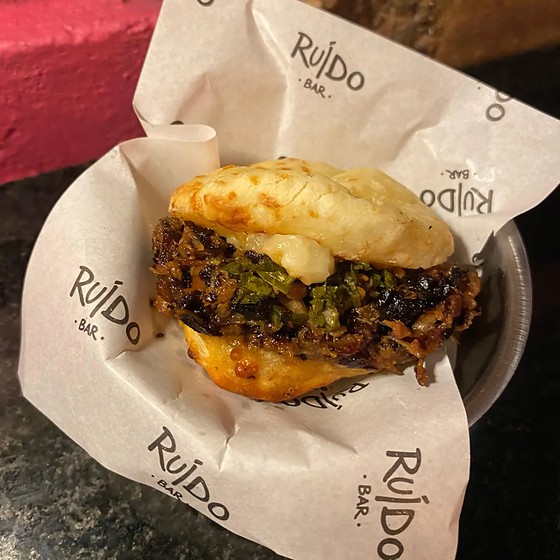 Pão de queijo recheado com porco desfiado e chimichurri, segurado na mão em frente a uma parede de folhagem.