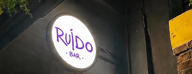 A placa redonda iluminada do Ruído Bar acesa na fachada preta, à noite, com folhagem pendurada por cima.